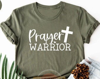 May include: Heather olive green t-shirt with the words "Prayer Warrior" and a white cross graphic. The shirt is folded, with a pair of leopard print shoes and a scrunchie, and light blue jeans.
