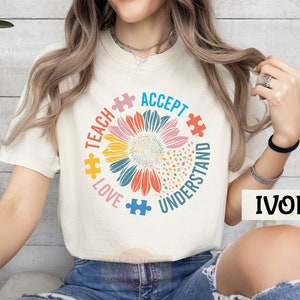 May include: Ivory-colored t-shirt featuring a colorful sunflower design with puzzle pieces and the words "Teach, Love, Accept, Understand." The shirt has short sleeves and a crew neck.