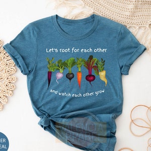 May include: A teal blue t-shirt with the text "Let's root for each other and watch each other grow" printed in white. The shirt features a graphic of five colorful vegetables, including a carrot, a turnip, a beet, a radish, and a parsnip.