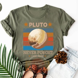 May include: A green t-shirt with a retro-style graphic of Pluto, the planet. The graphic includes a stylized image of Pluto with a red, orange, yellow, and blue striped background. The text "Pluto" is above the graphic, and the text "Never Forget 1930-2006" is below the graphic.