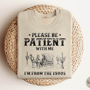 May include: Beige sweatshirt with a black graphic of a horse-drawn wagon and cacti. The text "Please be patient with me I'm from the 1900s" is printed on the sweatshirt.