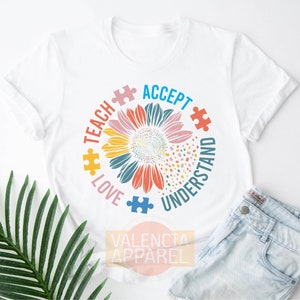 May include: White t-shirt with a colorful sunflower design and the words "Teach Love Accept Understand" around it. The sunflower is made up of puzzle pieces.