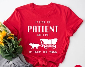 May include: Red t-shirt with white text that reads "Please be patient with me I'm from the 1900s". The text is in a pixelated font and there is a pixelated image of a covered wagon pulled by an ox.