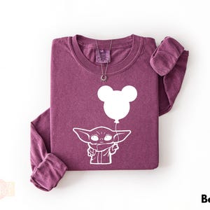 A purple long-sleeved t-shirt with a white graphic design depicting a young alien character holding a balloon.