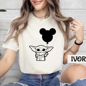 A woman wearing a white t-shirt with a graphic of a baby alien character holding a balloon, sitting against a white background.