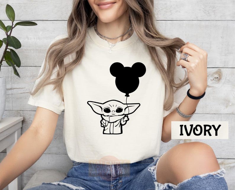 A woman wearing a white t-shirt with a graphic of a baby alien character holding a balloon, sitting against a white background.