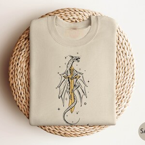 May include: A beige sweatshirt with a graphic print of a dragon wrapped around a sword. The dragon is outlined in black and the sword is gold. The background is a light beige color.