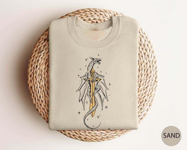 May include: A beige sweatshirt with a graphic print of a dragon wrapped around a sword. The dragon is outlined in black and the sword is gold. The background is a light beige color.