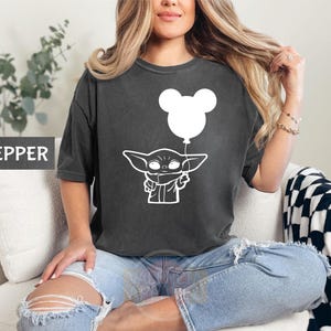 A woman wearing a black t-shirt with a white graphic of a baby Yoda character holding a balloon, sitting on a couch.