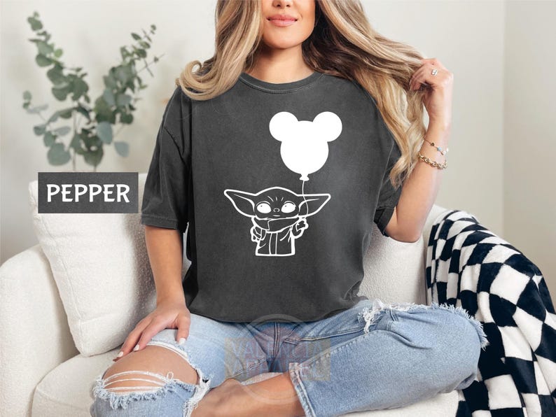 A woman wearing a black t-shirt with a white graphic of a baby Yoda character holding a balloon, sitting on a couch.