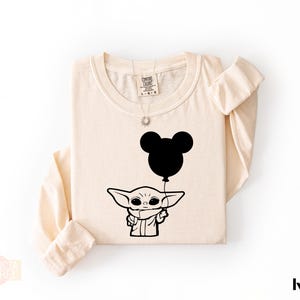 A beige t-shirt with a graphic design featuring a cartoon character holding a balloon with a Mickey Mouse face.