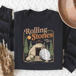 May include: Black crewneck sweatshirt with a graphic design featuring the text "Rolling Stones" above an illustration of a tomb with a stone and crosses, with the text "Since AD 33".