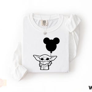 A white long-sleeved t-shirt with a black graphic design depicting a young alien character holding a balloon.