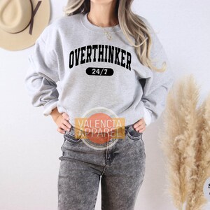 May include: A light gray sweatshirt with the word "OVERTHINKER" in black arched text above "24/7" in a black rectangle. The sweatshirt is paired with gray jeans. The image also includes a tan hat and dried ornamental grass.