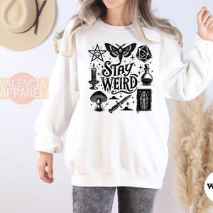 a woman wearing a white sweatshirt with the words stay weird on it