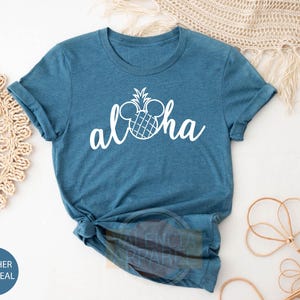 May include: A teal blue t-shirt with a white pineapple design and the word "Aloha" in a white cursive font. The pineapple design incorporates Mickey Mouse ears.