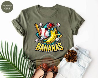 May include: Heather olive green t-shirt featuring a cartoon banana wearing sunglasses and a baseball cap, holding a bat, and the word "BANANAS". Baseball theme with baseballs in the background.