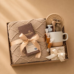 May include: A gift box containing a tan textured throw blanket with a diamond pattern, a Moonstruck dark chocolate espresso bar with a satin ribbon, a speckled ceramic mug, a small jar of honey with a wooden dipper, and a small gift-wrapped box.