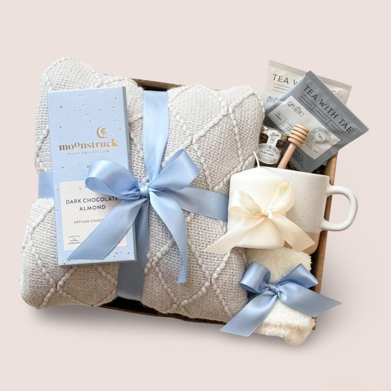 K&ouml;nnte beinhalten: Ein Geschenkkorb mit einer hellblauen Decke, einer Schachtel dunkler Schokoladenmandeln, einer wei&szlig;en Tasse, Teebeuteln, einer kleinen Kerze und einem Honigl&ouml;ffel. Die Artikel sind mit hellblauen und cremefarbenen B&auml;ndern gebunden. Die Decke hat ein Rautenmuster.