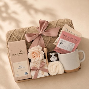 May include: A gift set containing a tan cable-knit blanket tied with a dusty rose ribbon, a Moonstruck milk chocolate bar, two Tea With Tae tea bags, a small jar of honey with a wooden dipper, a white ceramic mug, and a small white gift box.
