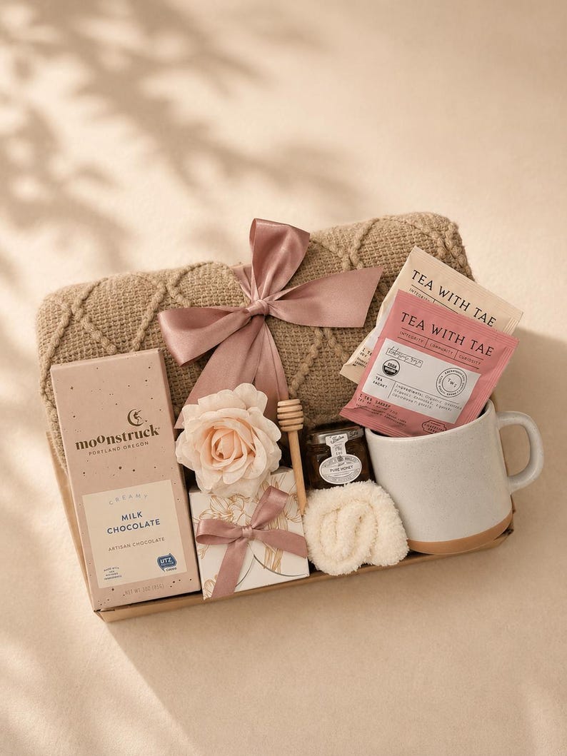 May include: A gift set containing a tan cable-knit blanket tied with a dusty rose ribbon, a Moonstruck milk chocolate bar, two Tea With Tae tea bags, a small jar of honey with a wooden dipper, a white ceramic mug, and a small white gift box.