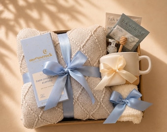May include: A gift box containing a beige textured knit blanket tied with a light blue ribbon, a Moonstruck dark chocolate almond bar, a ceramic mug with a cream ribbon, a small honey jar, a wooden honey dipper, tea packets, and a white washcloth.