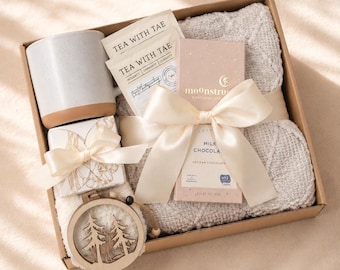 Op de afbeelding: Een geschenkdoos met een lichtgrijze keramische mok, twee Tea With Tae theezakjes, een Moonstruck melkchocoladereep met een crèmekleurig lint, een klein cadeaudoosje met bloemenprint en crèmekleurig lint, en een ronde houten hanger met bomen.