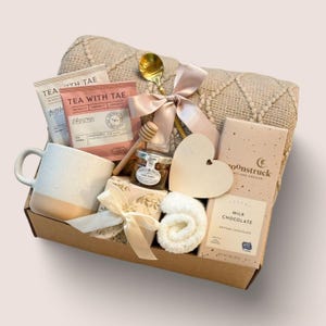 May include: A gift basket filled with a beige blanket, a white and peach-colored mug, tea packets, a jar of honey, a wooden heart, a bar of chocolate, and a small rolled washcloth. A gold spoon and a pink ribbon add decorative touches.