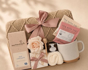 Op de afbeelding: Een geschenkset met een beige kabelgebreide deken vastgebonden met een oudroze lint, een Moonstruck melkchocoladereep, twee Tea With Tae theezakjes, een klein potje honing met een houten honingdipper, een witte keramische mok en een klein wit geschenkdoosje.