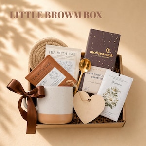 May include: A gift box labeled LITTLE BROWM BOX containing a white and tan speckled mug with a brown ribbon, a round woven coaster, two Tea With Tae packets, a Moonstruck dark chocolate bar, a gold spoon, a wooden heart tag, and a Gardenia sachet.