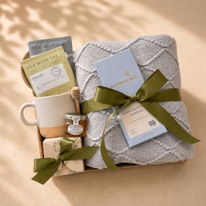 May include: A gift set containing a light gray textured knit blanket, a speckled ceramic mug, a small jar of honey with a wooden dipper, a box of Tea With Tae tea bags, and a Moonstruck dark chocolate almond bar, all tied with olive green ribbons.