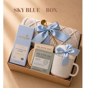 May include: A gift box containing a light blue Moonstruck dark chocolate almond bar, a textured beige blanket, a gold spoon, two Tea With Tae tea sachets, and a white ceramic mug with a light blue satin bow. The text SKY BLUE BOX is visible.