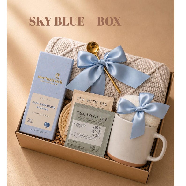 May include: A gift box containing a light blue Moonstruck dark chocolate almond bar, a textured beige blanket, a gold spoon, two Tea With Tae tea sachets, and a white ceramic mug with a light blue satin bow. The text SKY BLUE BOX is visible.