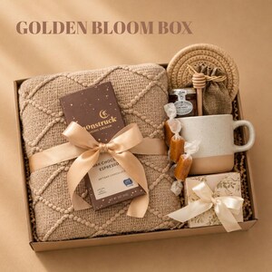 May include: A gift box labeled GOLDEN BLOOM BOX containing a tan textured throw blanket, a Moonstruck dark chocolate espresso bar with a gold ribbon, a speckled ceramic mug, a small jar of honey, a wooden honey dipper, and a small floral gift box.