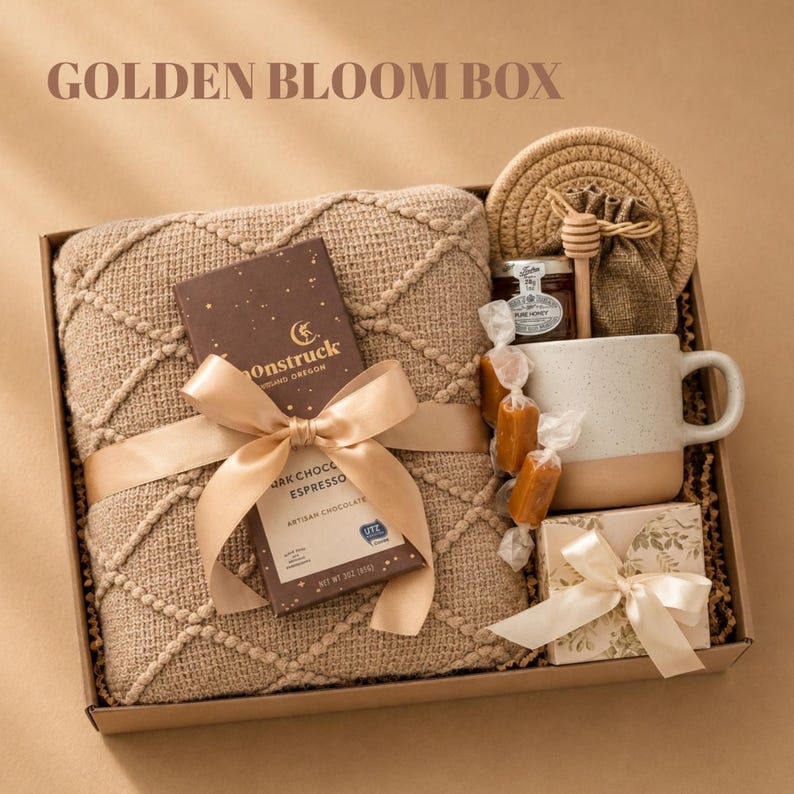 May include: A gift box labeled GOLDEN BLOOM BOX containing a tan textured throw blanket, a Moonstruck dark chocolate espresso bar with a gold ribbon, a speckled ceramic mug, a small jar of honey, a wooden honey dipper, and a small floral gift box.