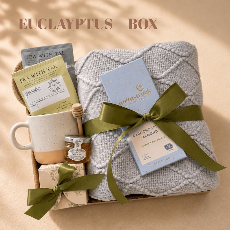 May include: A gift box containing a light gray textured throw blanket tied with an olive green ribbon, a Moonstruck dark chocolate almond bar, a speckled ceramic mug, a small jar of honey with a wooden dipper, and Tea With Tae tea packets.