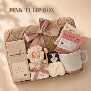 May include: A gift box labeled PINK TULIP BOX containing a tan textured blanket with a pink satin ribbon, a Moonstruck milk chocolate bar, a pink rose, a wooden honey dipper, a small jar of honey, a white mug, and Tea With Tae tea packets.