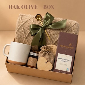 May include: A gift box labeled OAK OLIVE BOX containing a beige textured knit blanket with a green ribbon and gold spoon, a white ceramic mug, a small jar, a burlap pouch, a wooden heart, and a Moonstruck Dark Chocolate Espresso bar.