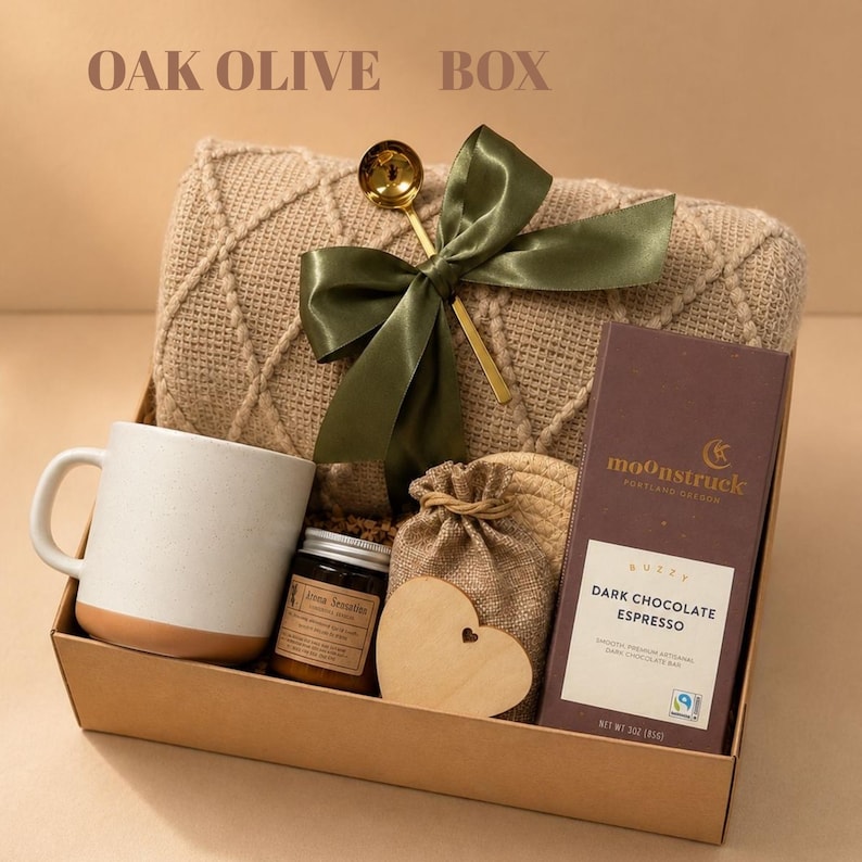 May include: A gift box labeled OAK OLIVE BOX containing a beige textured knit blanket with a green ribbon and gold spoon, a white ceramic mug, a small jar, a burlap pouch, a wooden heart, and a Moonstruck Dark Chocolate Espresso bar.