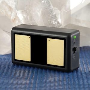 May include: A black rectangular device featuring two beige panels and a black central section. The device has a green light, a switch, and two ports on the side. It rests on a blue textured surface.