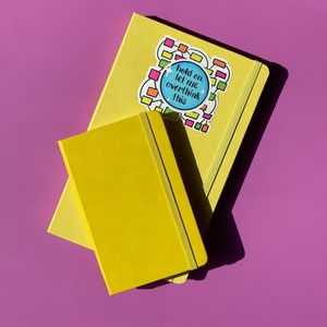 May include: Two yellow notebooks stacked on top of each other. The top notebook has a sticker on it that says "Hold on, let me overthink this". The sticker is a blue circle with white text and a colorful design around it.