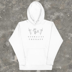 May include: White hoodie with a black floral design and the text "NORMALIZE THERAPY" printed on the front.
