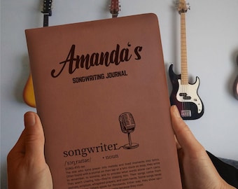 May include: A brown songwriting journal titled "Amanda's SONGWRITING JOURNAL" with a microphone graphic. The journal includes the definition of "songwriter". Guitars hang in the background. The journal is held by a person.