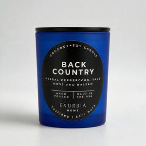 Puede incluir: Una vela en frasco de vidrio esmerilado azul con tapa negra y etiqueta circular negra. La etiqueta dice COCONUT+SOY CANDLE BACK COUNTRY HERBAL PEPPERCORN, SAGE MOSS AND BALSAM HAND POURED MADE IN THE USA EXURBIA HOME 7 oz/198 g | 50 hr burn.