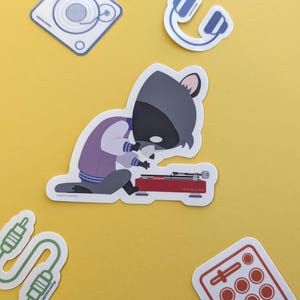 May include: A collection of stickers on a yellow background. The stickers feature a cartoon raccoon DJ, headphones, audio cables, a mixing board, and a speaker. The raccoon is wearing a purple jacket and is operating a red turntable.