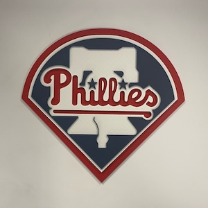 May include: A red, white, and blue baseball team logo with the word "Phillies" in a white, red, and blue script font. The logo is in the shape of a diamond with a Liberty Bell inside.