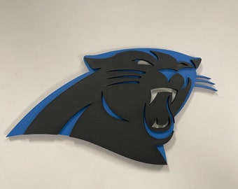 May include: A black and blue Carolina Panthers logo, a stylized panther head with an open mouth and visible teeth. The logo is layered, with a black panther shape over a blue outline.
