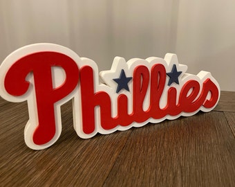 May include: White and red 3D sign with the word "Phillies" in red letters with two blue stars above the "i".