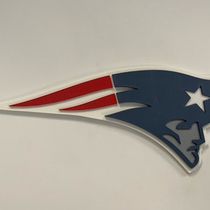 May include: A white, blue, and red 3D wall decor featuring the New England Patriots logo. The logo is a stylized silhouette of a patriot's head with a star above it. The logo is outlined in white and has a red stripe across the top.