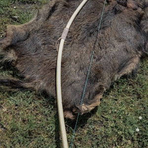 May include: A wooden bow with a string attached, lying on a brown animal hide. The bow is made of natural wood and has a simple design.